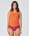 Lächelnde Frau in orangem Trägertop mit feinem Muster und rotem Slip mit „Pappenberg“-Aufdruck, stehend vor neutralem, hellem Hintergrund.