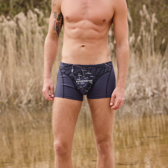 Mann trägt dunkelblaue Boxershorts mit weißem Aufdruck Pulverhörndl und Bergmotiv von Ein schöner Fleck Erde, im Hintergrund Schilf und Wasser