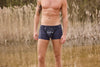 Mann trägt dunkelblaue Boxershorts mit weißem Aufdruck Pulverhörndl und Bergmotiv von Ein schöner Fleck Erde, im Hintergrund Schilf und Wasser