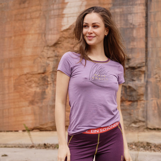 Frau trägt lilafarbenes T-Shirt mit Aufdruck Wald knallt und violette Funktionsleggings mit rotem Bund von Ein schöner Fleck Erde, Outdooraufnahme vor Felswand