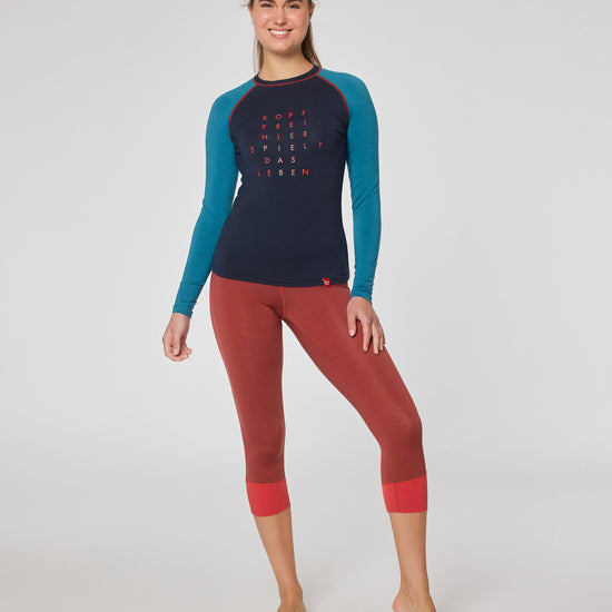 Frau trägt ein dunkelblaues Longsleeve mit türkisfarbenen Ärmeln und eine rote 7/8-Leggings, steht barfuß im Studio und lächelt in die Kamera.
