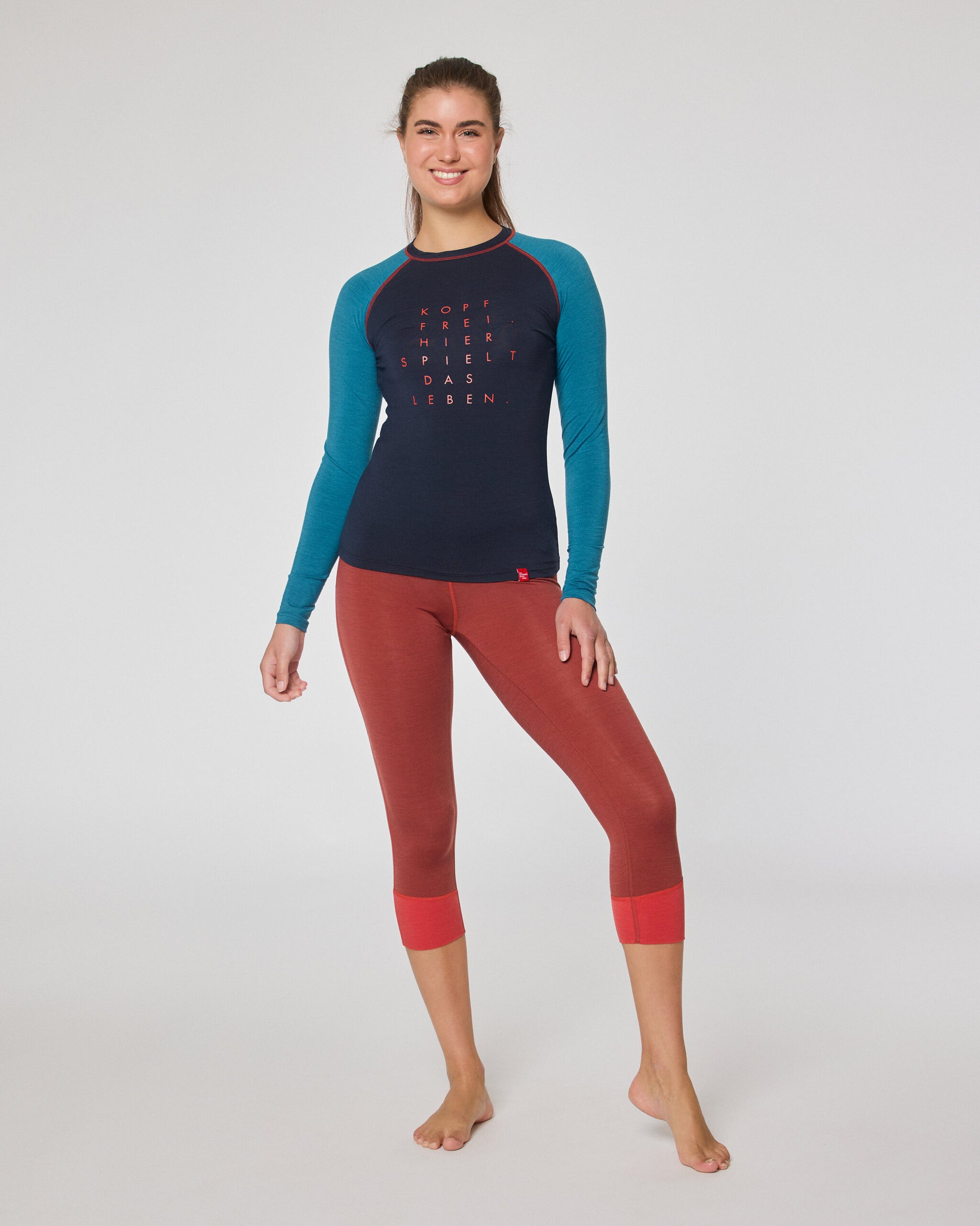 Frau trägt ein dunkelblaues Longsleeve mit türkisfarbenen Ärmeln und eine rote 7/8-Leggings, steht barfuß im Studio und lächelt in die Kamera.