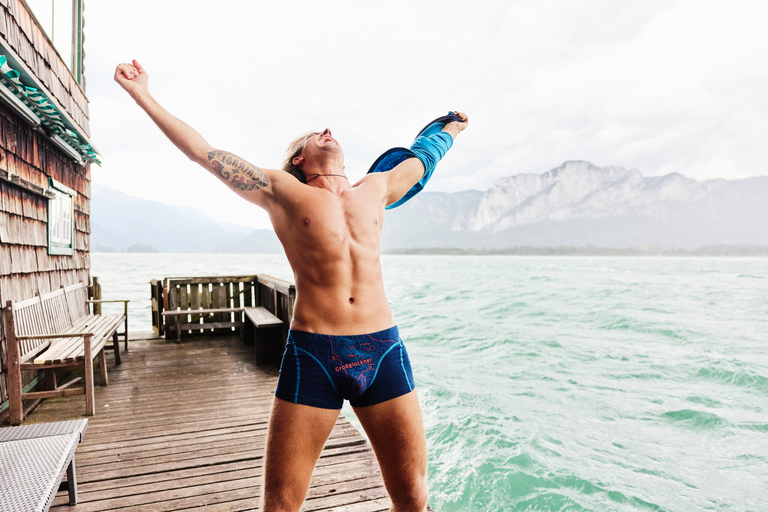 Mann in blauer Boxershorts von Ein schöner Fleck Erde mit rotem Aufdruck ‚Großglockner‘, stehend auf einem Steg am See, streckt jubelnd die Arme in die Höhe, im Hintergrund Berge und Wasser.