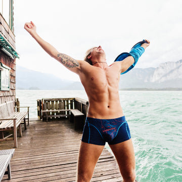 Mann steht auf einem Holzsteg am Wasser und trägt eine dunkelblaue Herren-Boxershorts mit rotem topografischem Linienmuster und weiß-rotem Schriftzug ‚Großglockner‘; im Hintergrund sind See und Berge zu sehen.