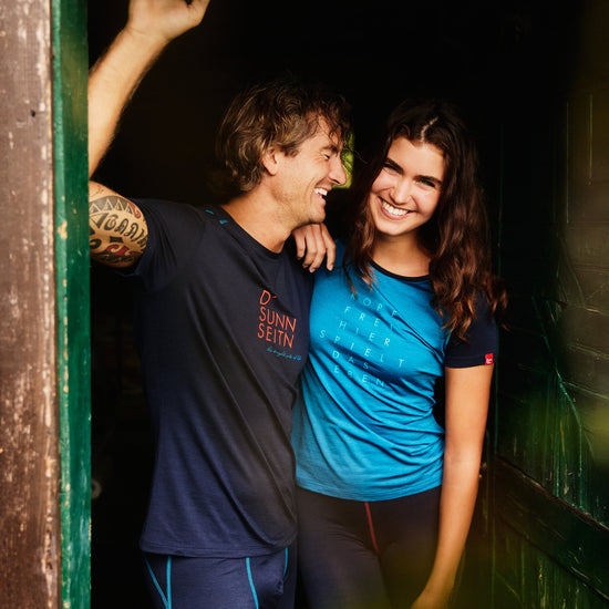 Mann und Frau in Funktionskleidung von Ein schöner Fleck Erde, lachend im Türrahmen einer Holzhütte – beide tragen Shirts mit Schriftzug ‚D’ Sunn Seitn‘.