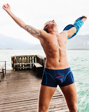 Mann in dunkelblauer Boxershorts mit rotem Aufdruck ‚Gipfelstürmer‘, stehend auf einem Holzsteg am See, reißt jubelnd die Arme hoch und zieht ein Shirt aus, Berge und Wasser im Hintergrund