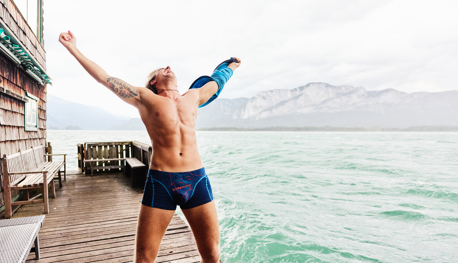 Mann in dunkelblauer Boxershorts mit rotem Aufdruck ‚Großglockner‘, stehend mit ausgebreiteten Armen auf einem Holzsteg am See mit Bergen im Hintergrund