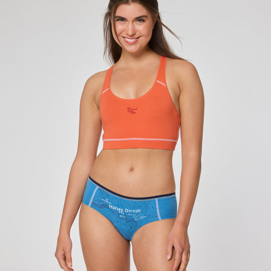Lächelnde Frau in orangefarbenem Sport-BH und blauer Hipster-Unterhose mit topografischem Kartendruck. Auf der Unterhose steht die Aufschrift „Hohes Dirndl“.