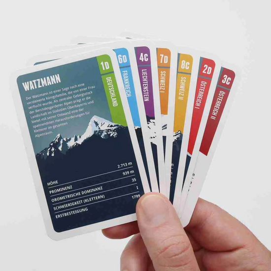 Hand hält Spielkarten des Alpen-Quartetts von Ein schöner Fleck Erde, mit Bergmotiven wie dem Watzmann und Angaben zu Höhe, Prominenz und Kletterschwierigkeit – inspiriert von den Alpenmotiven der Unterwäschekollektion.