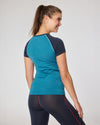 Frau in pfauenblauem Kurzarm-Funktionsshirt mit dunkelblauen Raglanärmeln von Ein schöner Fleck Erde, Rückenansicht vor grauem Hintergrund, dazu dunkle Leggings mit roter Naht.