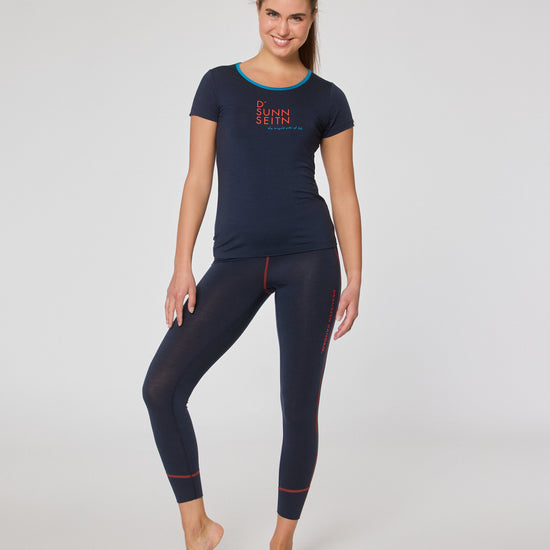 Frau trägt ein dunkelblaues Kurzarmshirt mit Print und eine dunkelblaue Leggings mit roten Nähten, steht barfuß leicht seitlich lächelnd vor der Kamera.
