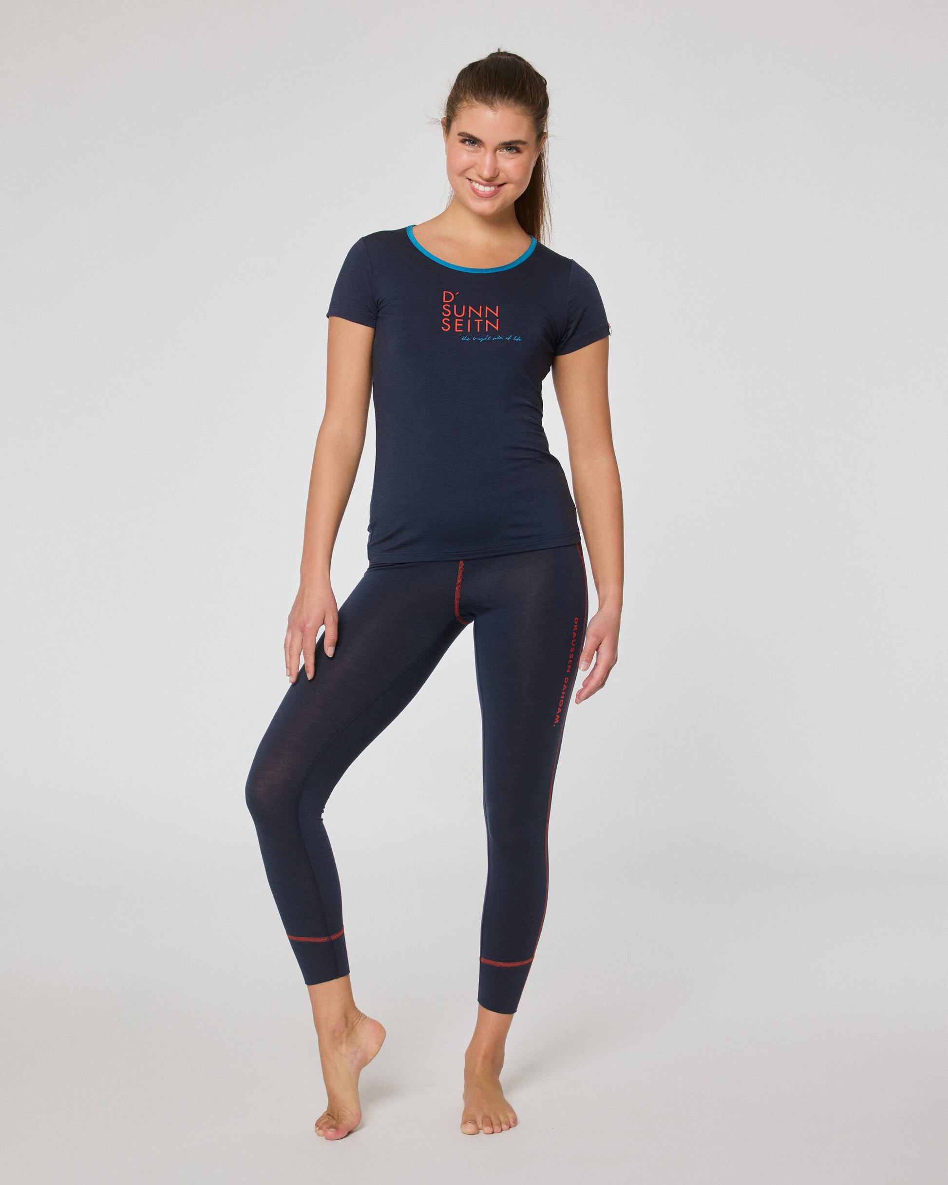 Frau trägt ein dunkelblaues Kurzarmshirt mit Print und eine dunkelblaue Leggings mit roten Nähten, steht barfuß leicht seitlich lächelnd vor der Kamera.