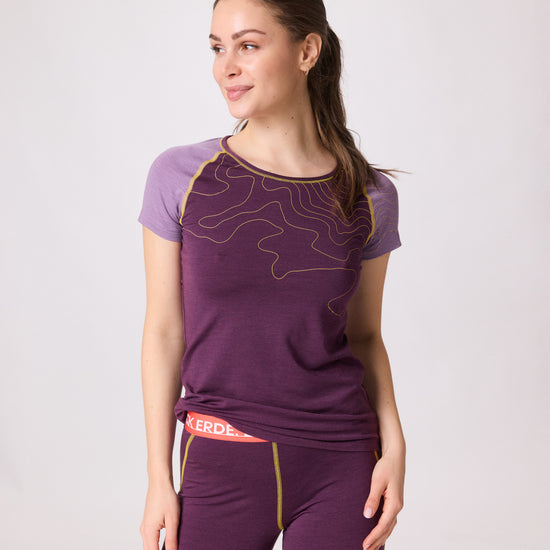 Frau trägt violettes Funktionsshirt mit gelben Linien und passende ¾-Leggings mit rotem Bund von Ein schöner Fleck Erde, Studioaufnahme mit hellem Hintergrund“