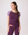 Frau trägt violettes Funktionsshirt mit gelben Linien und passende ¾-Leggings mit rotem Bund von Ein schöner Fleck Erde, Studioaufnahme mit hellem Hintergrund“