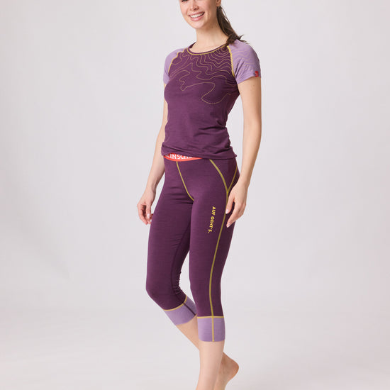 Frau trägt violettes Kurzarmshirt und passende 3/4-Funktionshose von Ein schöner Fleck Erde mit gelben Nähten und rotem Bund, steht seitlich und lächelt
