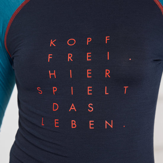 Nahaufnahme eines dunkelblauen Longsleeves mit türkisfarbenen Ärmeln und rotem Aufdruck auf der Brust: ‚KOPF FREI. HIER SPIELT DAS LEBEN.‘