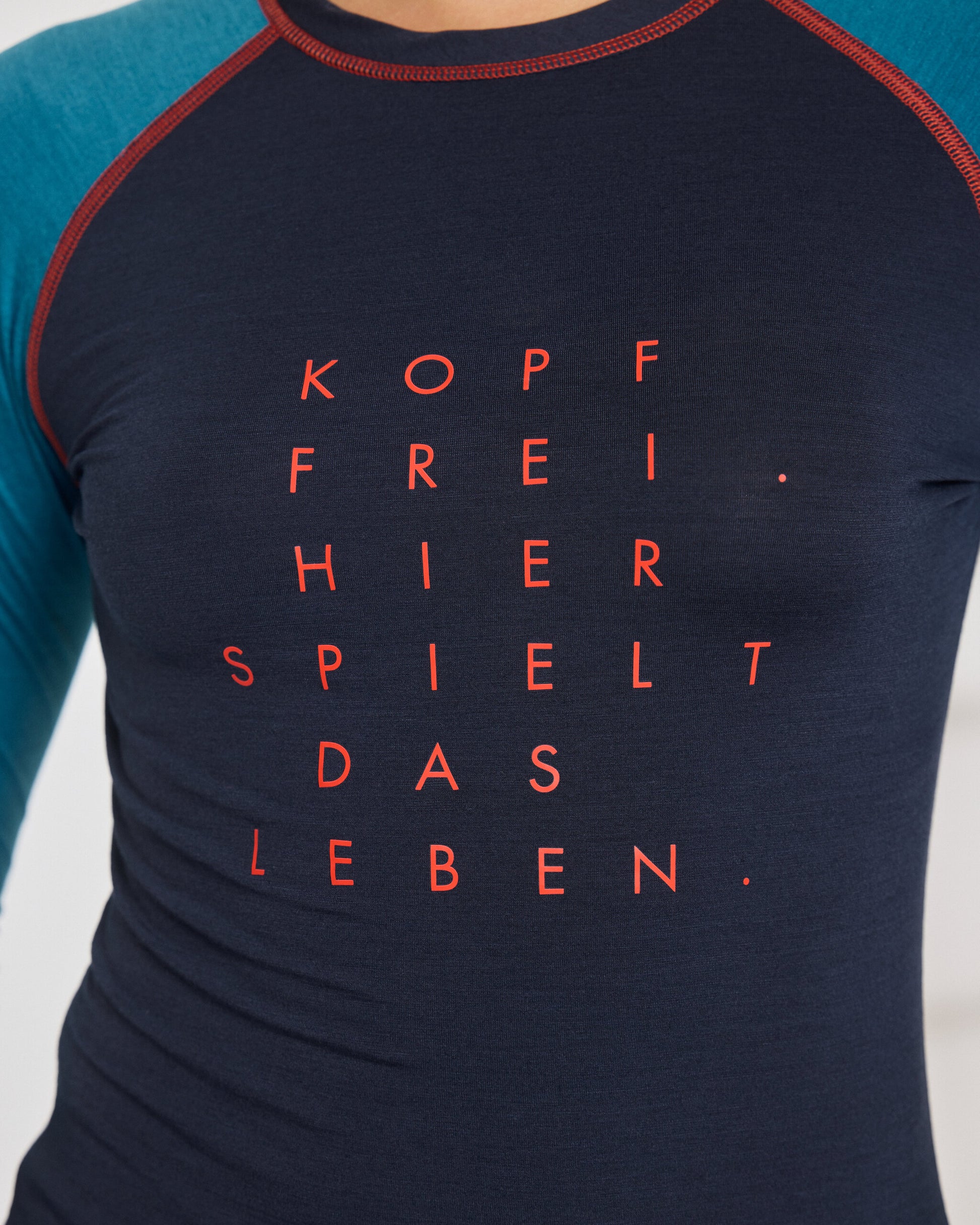 Nahaufnahme eines dunkelblauen Longsleeves mit türkisfarbenen Ärmeln und rotem Aufdruck auf der Brust: ‚KOPF FREI. HIER SPIELT DAS LEBEN.‘