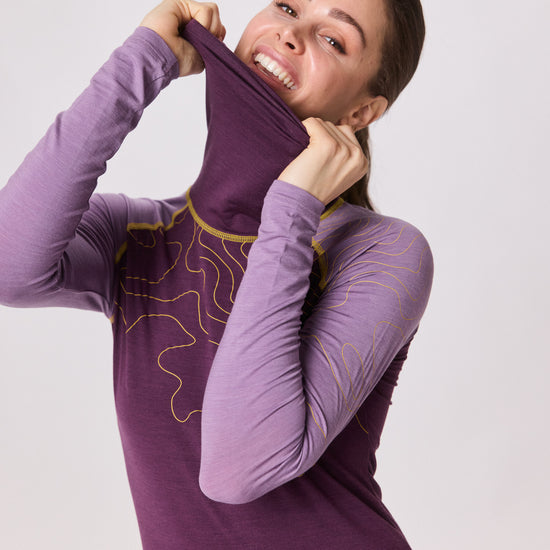 Frau trägt violettes Langarmshirt von Ein schöner Fleck Erde und zieht spielerisch den hohen Kragen über Mund und Nase, während sie lacht