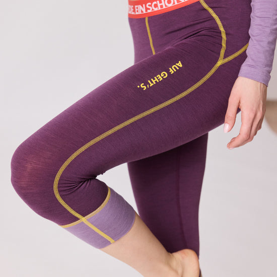 „Detailaufnahme der violetten 3/4-Funktionshose von Ein schöner Fleck Erde mit gelben Nähten, rotem Bund und Aufschrift ‚AUF GEHT’S‘ am Bein