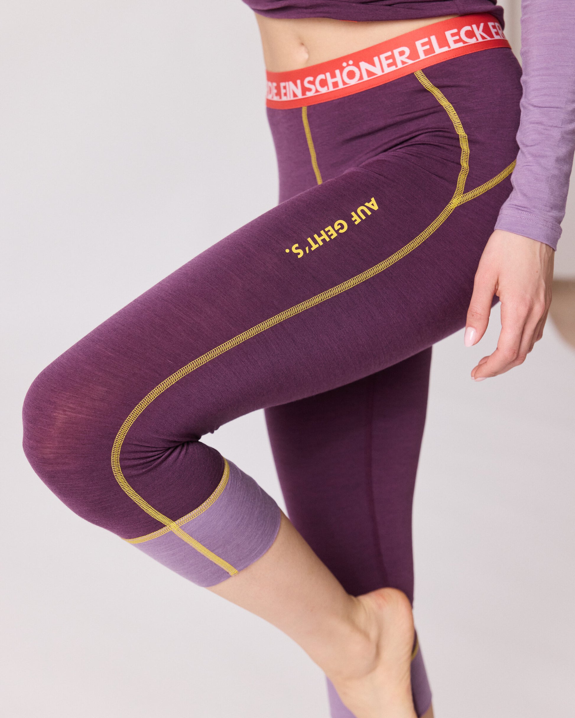 „Detailaufnahme der violetten 3/4-Funktionshose von Ein schöner Fleck Erde mit gelben Nähten, rotem Bund und Aufschrift ‚AUF GEHT’S‘ am Bein