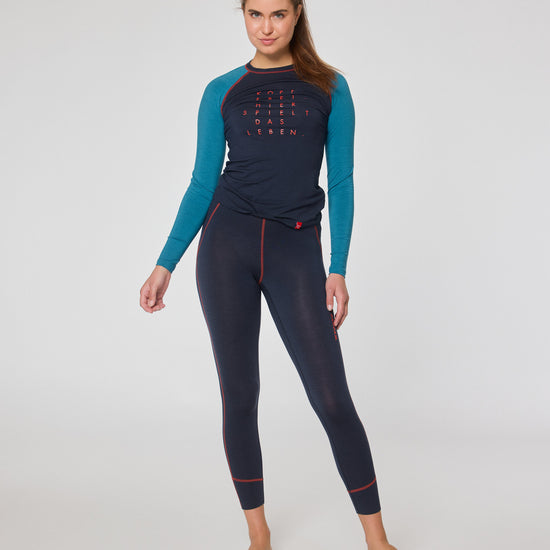 Frau trägt ein dunkelblaues Funktionslongsleeve mit türkisfarbenen Ärmeln und eine dunkelblaue Leggings mit roten Nähten, steht barfuß im Studio und blickt in die Kamera.