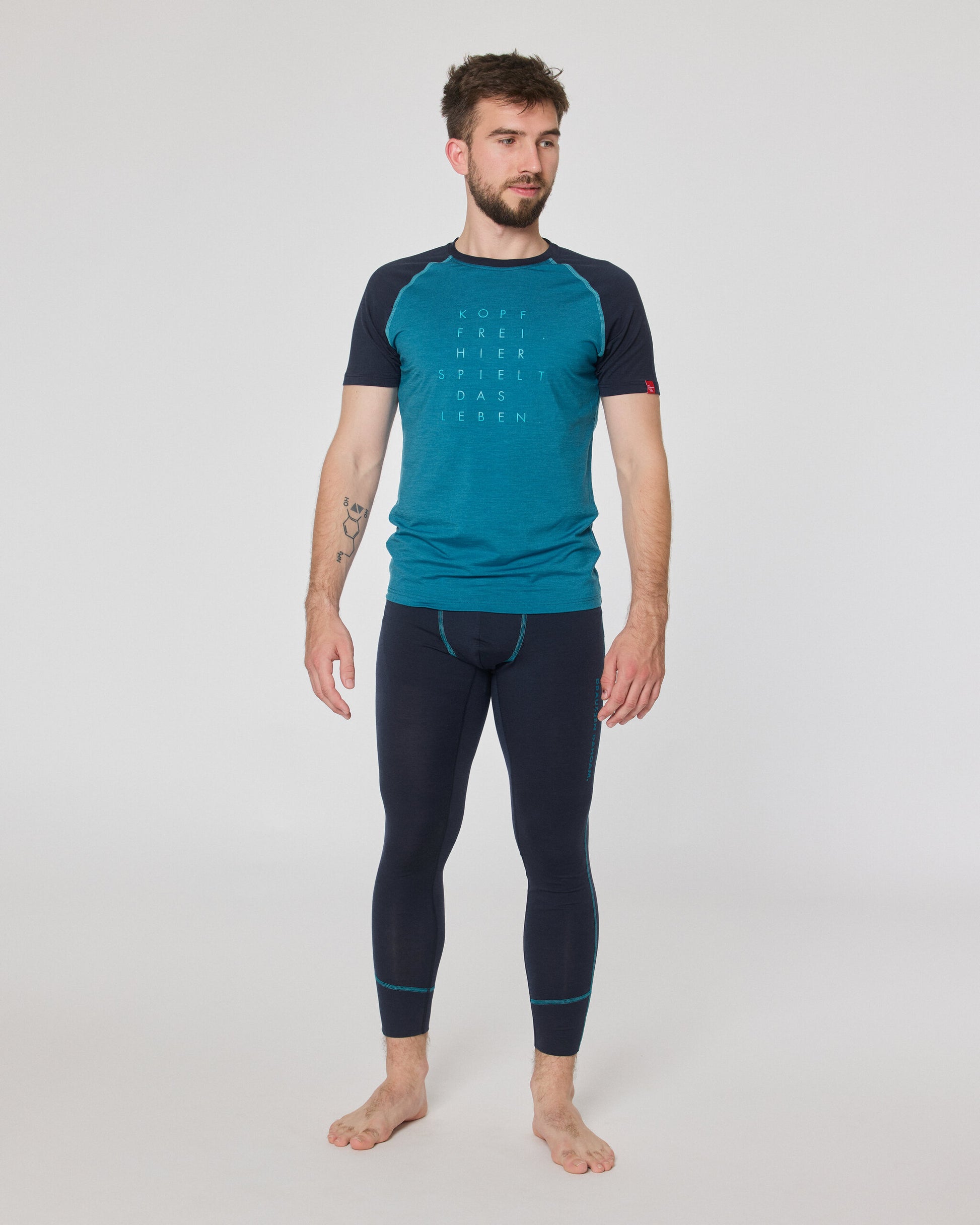 Mann trägt dunkelblaue Funktionsleggings und türkisfarbenes Kurzarmshirt von Ein schöner Fleck Erde, stehend vor neutralem Hintergrund