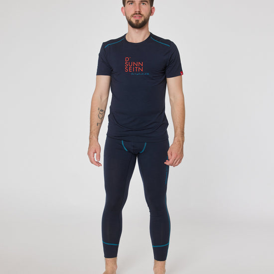 Mann trägt dunkelblaues Kurzarmshirt mit Aufdruck ‚D’ Sunnseitn‘ und passende dunkelblaue Funktionsleggings von Ein schöner Fleck Erde, stehend vor neutralem Hintergrund.