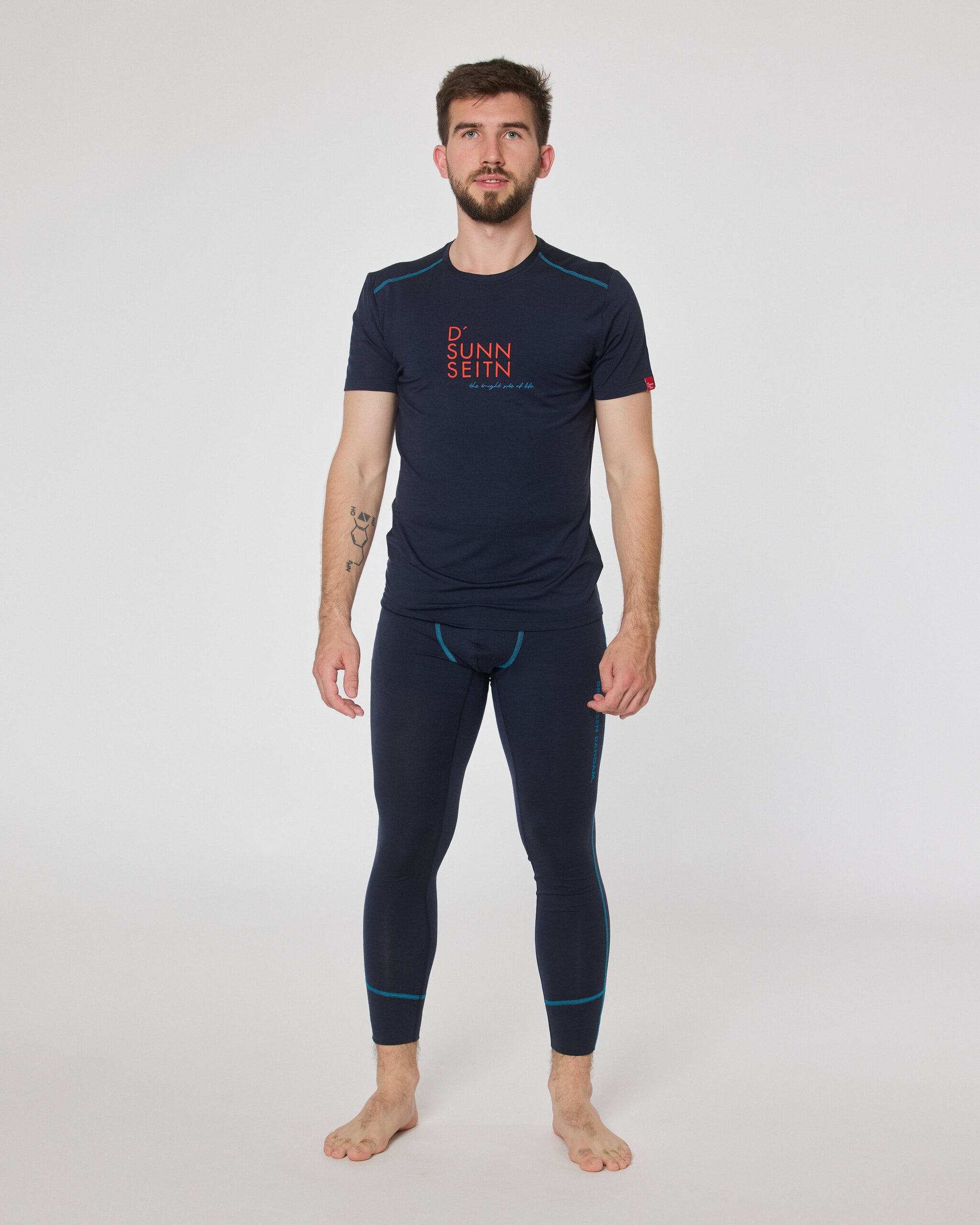 Mann trägt dunkelblaues Kurzarmshirt mit Aufdruck ‚D’ Sunnseitn‘ und passende dunkelblaue Funktionsleggings von Ein schöner Fleck Erde, stehend vor neutralem Hintergrund.