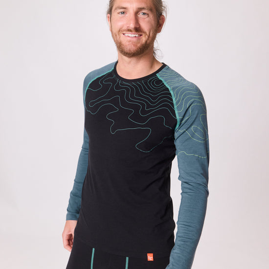 Mann trägt schwarzes Langarmshirt mit türkisfarbenen Ärmeln und Linienmuster von Ein schöner Fleck Erde, Studioaufnahme mit hellem Hintergrund
