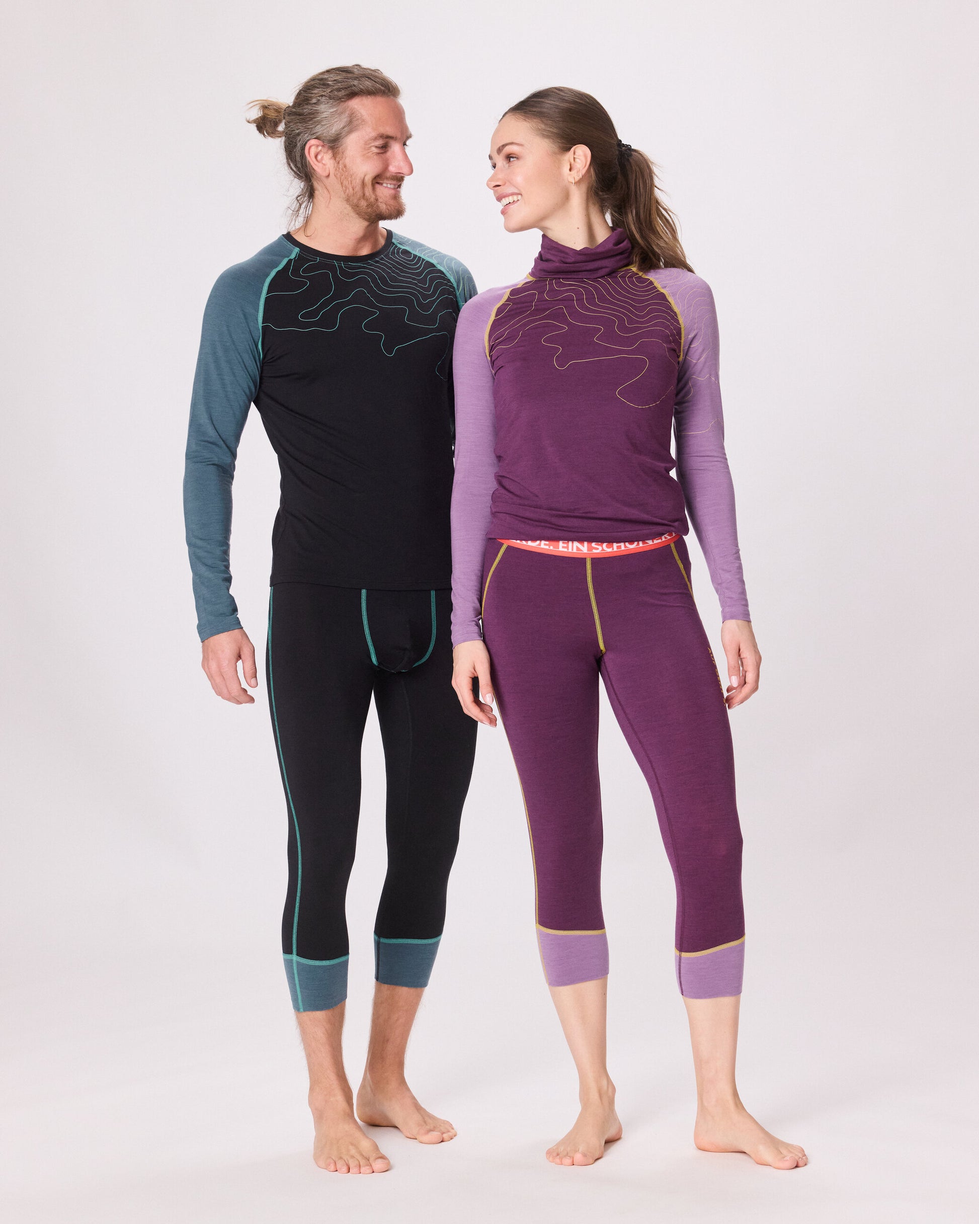 Ein Paar trägt passende Funktionswäsche von Ein schöner Fleck Erde: Er im dunkelblauen Langarmshirt mit hellblauen Ärmeln und sie im violetten Set mit gelben Nähten, gemeinsam im Studio fotografiert